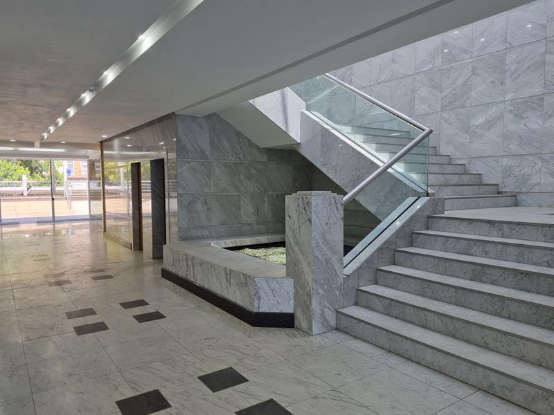 Spacious office building entrance lobby featuring high-end marble flooring and wall cladding, a modern glass staircase, and integrated elevator access.