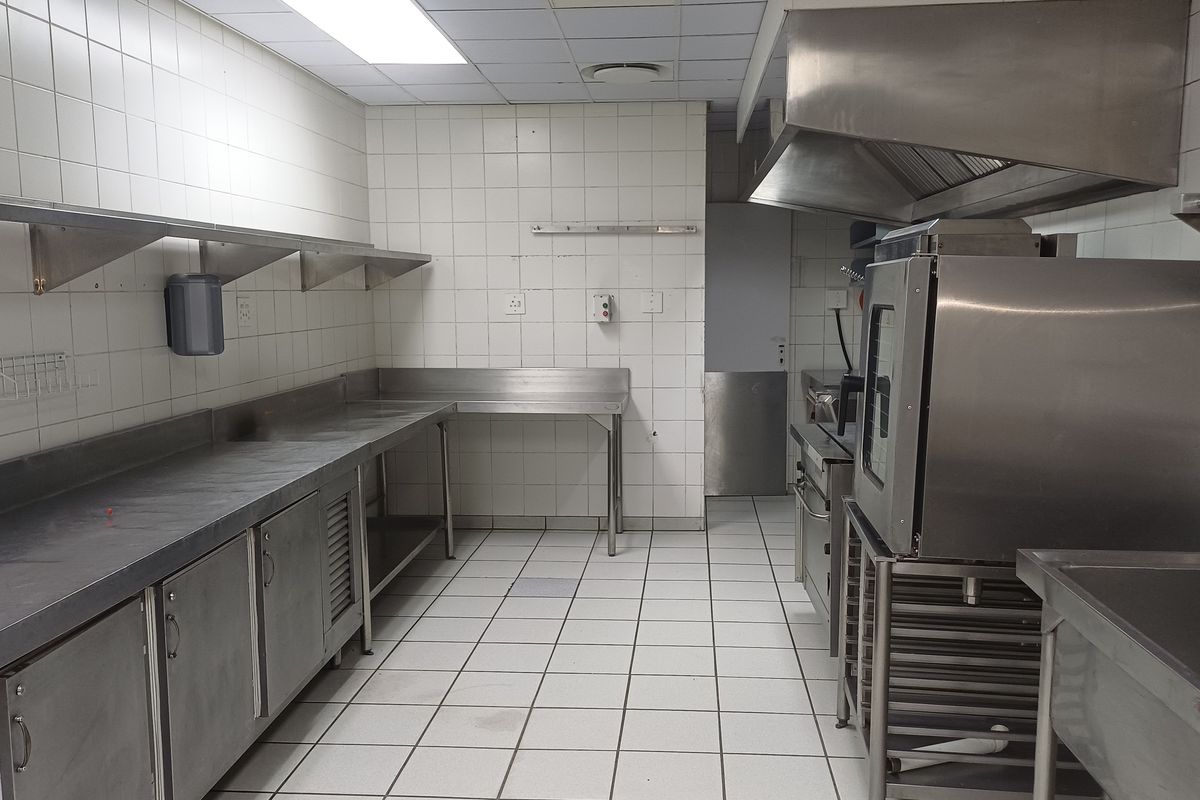 A functional commercial kitchen space featuring stainless steel work surfaces, integrated cabinetry, a professional extractor hood, and durable tiled finishes throughout.