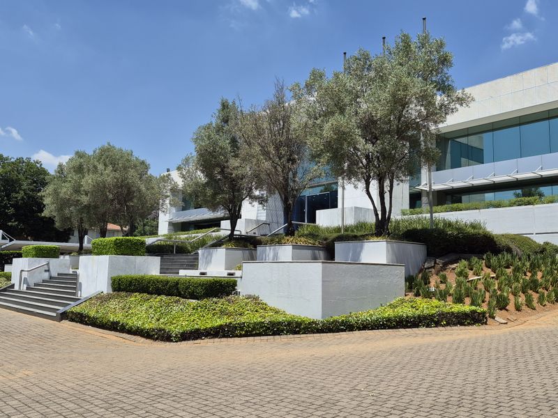 Modern office building exterior featuring a professional glass facade, tiered landscaped planters, and paved access pathways with concrete staircases.