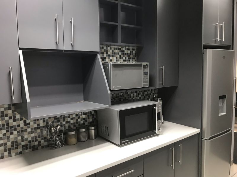 Modern office kitchen area featuring dark grey cabinetry, mosaic tile backsplash, and clean white countertops.