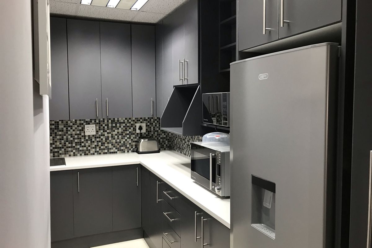 A modern office kitchenette featuring dark grey cabinetry, white countertops, a mosaic tile backsplash, and integrated lighting.