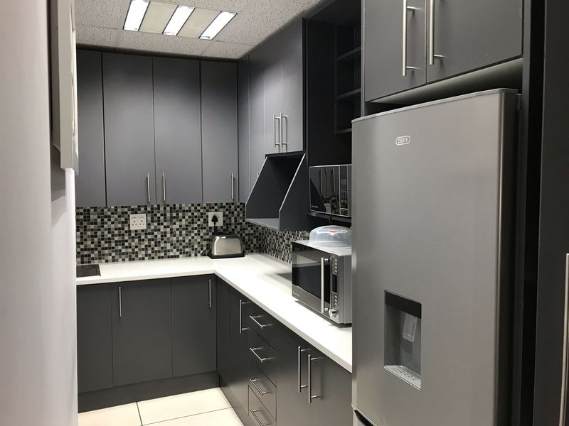 A modern office kitchenette featuring dark grey cabinetry, white countertops, a mosaic tile backsplash, and integrated lighting.