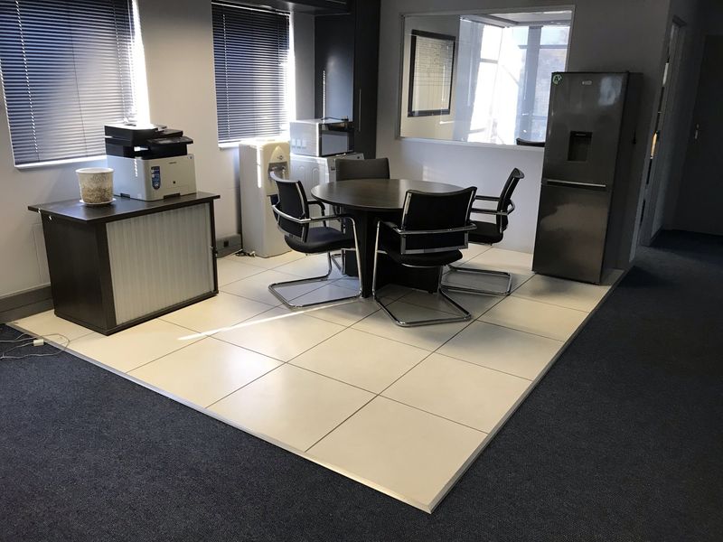 A functional office breakout area featuring a raised tiled floor, internal glass partitioning, and adjustable venetian blinds for light control.