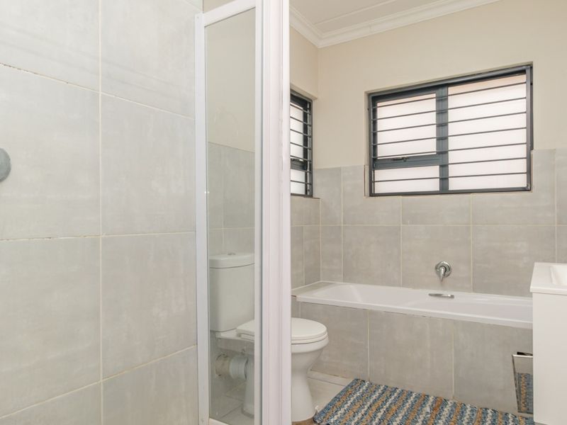 A clean, functional bathroom featuring a bathtub, shower, toilet, and vanity basin with neutral grey wall tiling and security bars on the window.