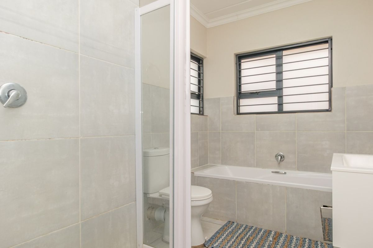 A clean, functional bathroom featuring a bathtub, shower, toilet, and vanity basin with neutral grey wall tiling and security bars on the window.