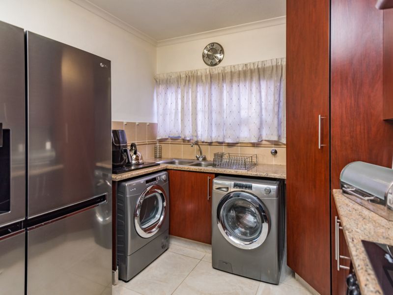 A functional scullery featuring granite countertops, double sink, tiled backsplash, and dedicated space for laundry appliances with ample built-in cabinetry.
