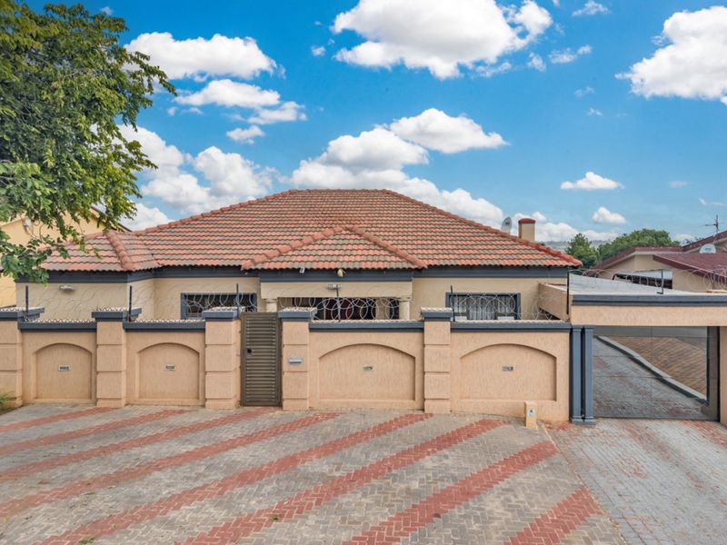Front elevation of a single-story house featuring a tiled roof, secure perimeter wall with razor wire, and a paved driveway with gated access.