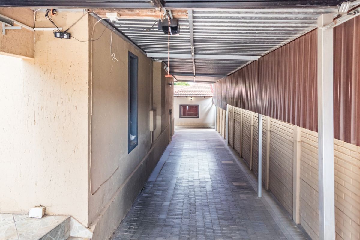 A narrow paved outdoor walkway covered by a corrugated metal roof, featuring boundary walls and exposed electrical infrastructure.