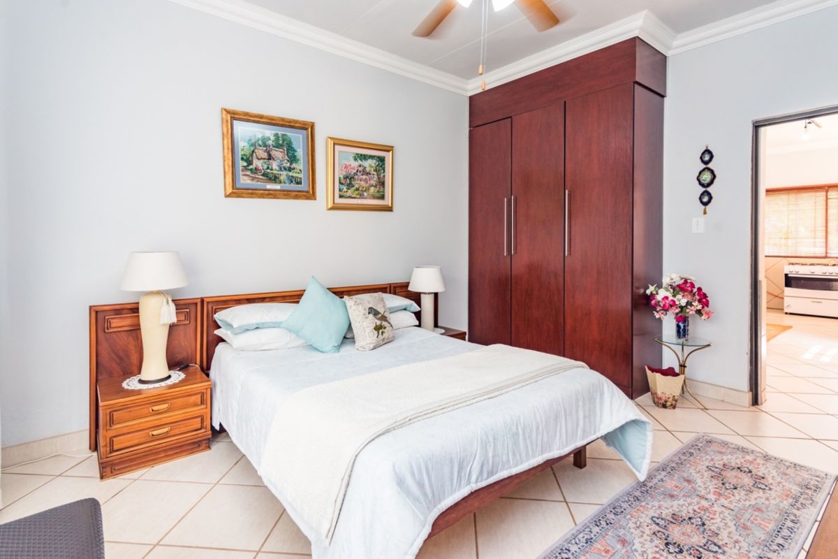 A well-maintained bedroom featuring built-in dark wood cabinetry, crown molding, and tiled flooring, with an adjacent view into a kitchen area.