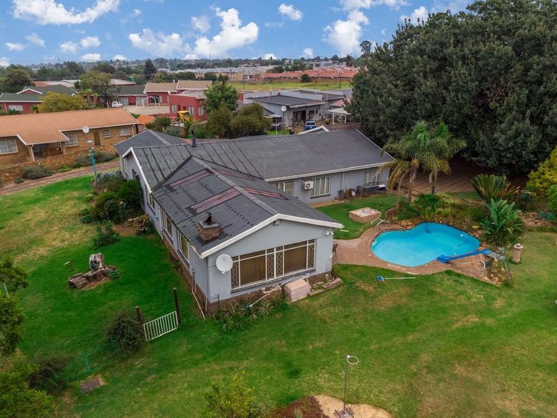 Aerial view of a small holding property featuring a residential dwelling with a metal roof, a swimming pool, and expansive garden grounds.