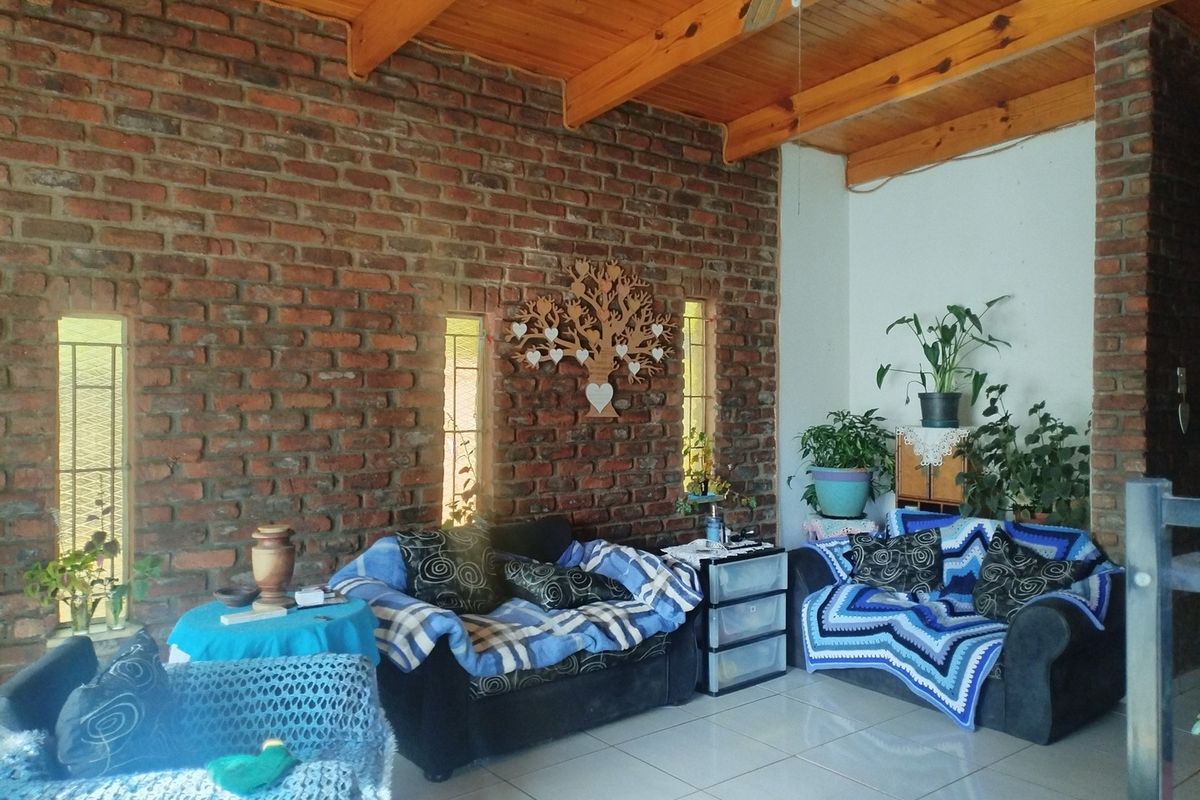 A cozy living room featuring an exposed brick feature wall, high timber beam ceilings, and tiled flooring with natural light from vertical windows.