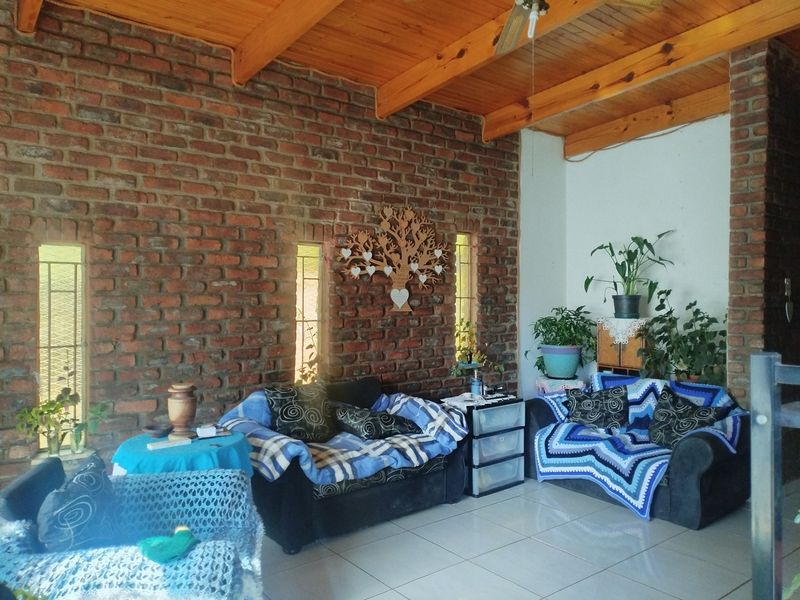 A cozy living room featuring an exposed brick feature wall, high timber beam ceilings, and tiled flooring with natural light from vertical windows.