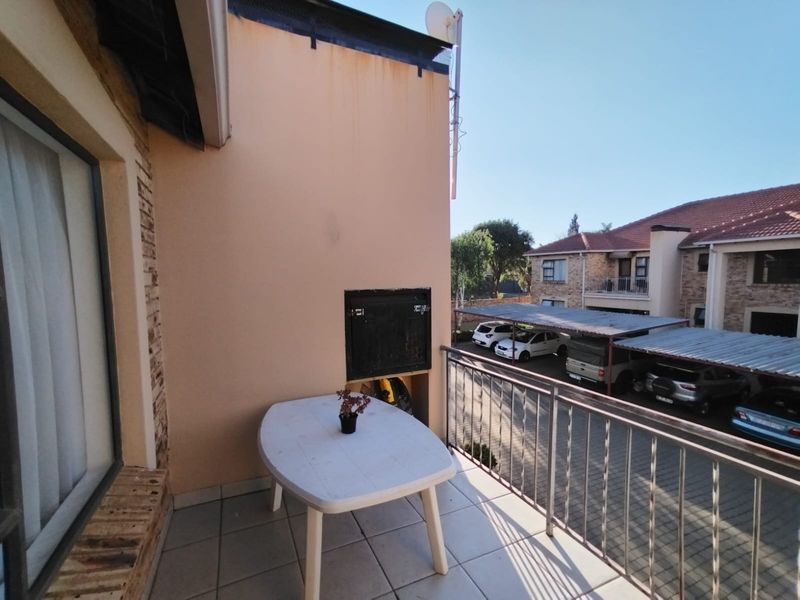 A balcony featuring tiled flooring, a built-in braai, and a metal balustrade, overlooking the apartment complex parking area.