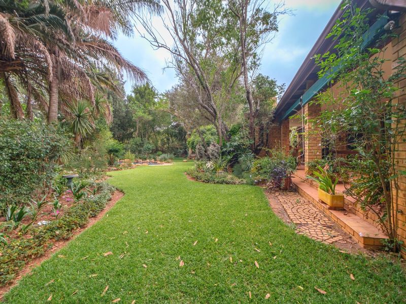 A lush, well-established garden featuring a manicured lawn, mature trees, and a covered brick patio area along the side of the house.