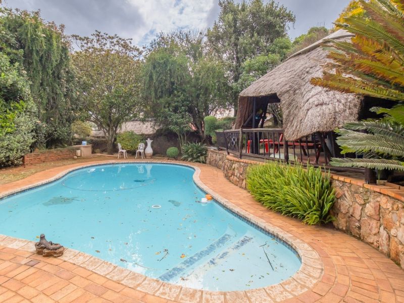 Outdoor pool area featuring a swimming pool, stone retaining wall, brick paving, and a traditional thatched lapa structure set within a garden.