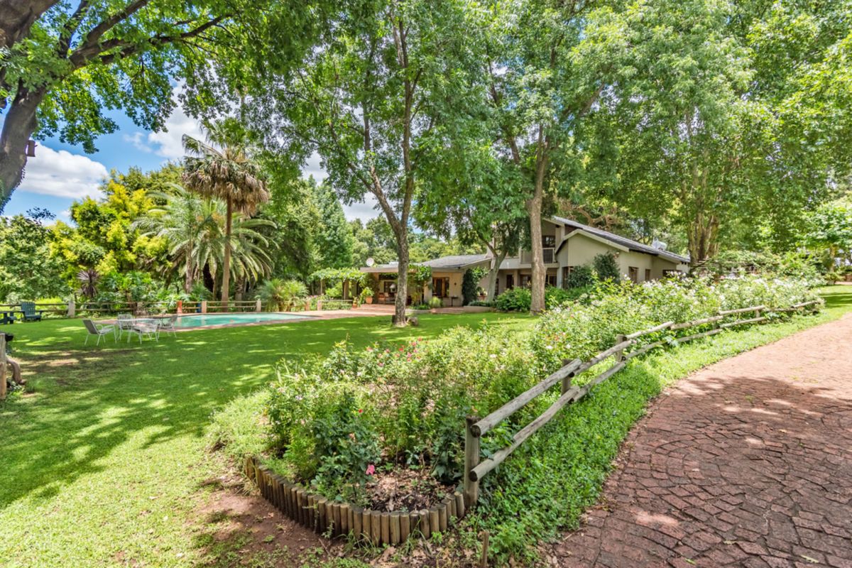 A scenic rear view of the farm property featuring a lush garden, mature trees, a swimming pool, and a paved driveway.
