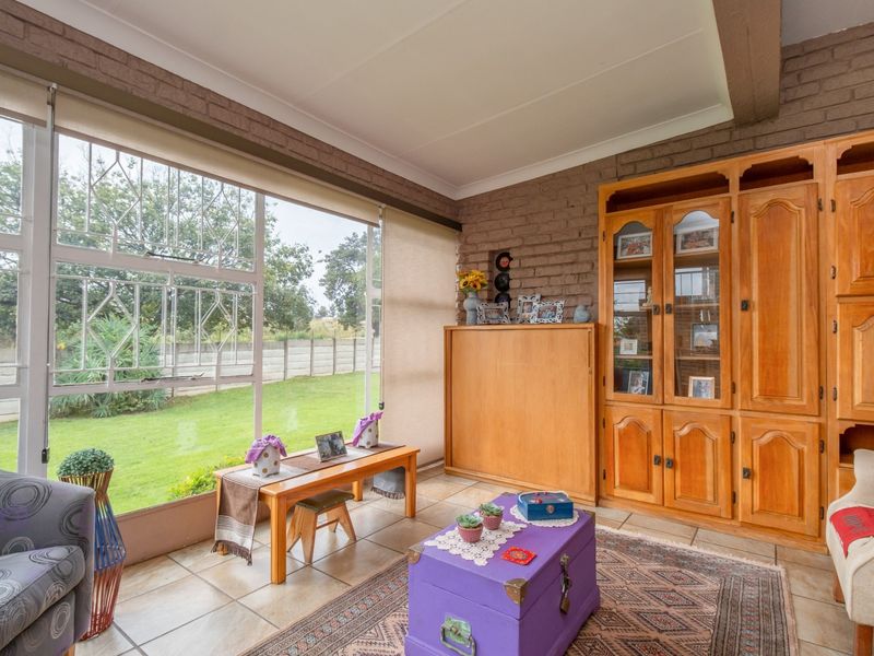 A sunlit living area featuring exposed brick walls, tiled flooring, and large windows with security bars, offering a view of the garden.