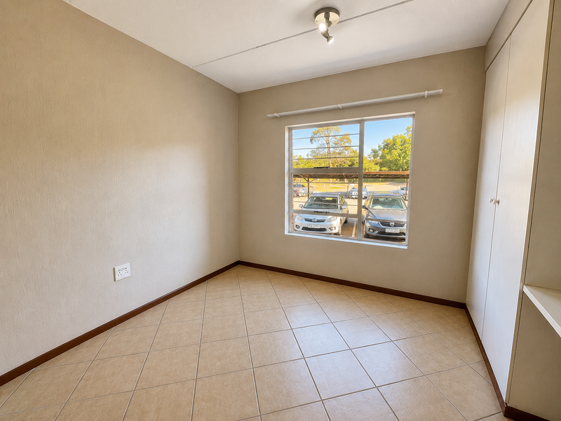 A bright, empty bedroom featuring neutral tiled flooring, built-in cupboards, and a window with security bars overlooking the parking area.