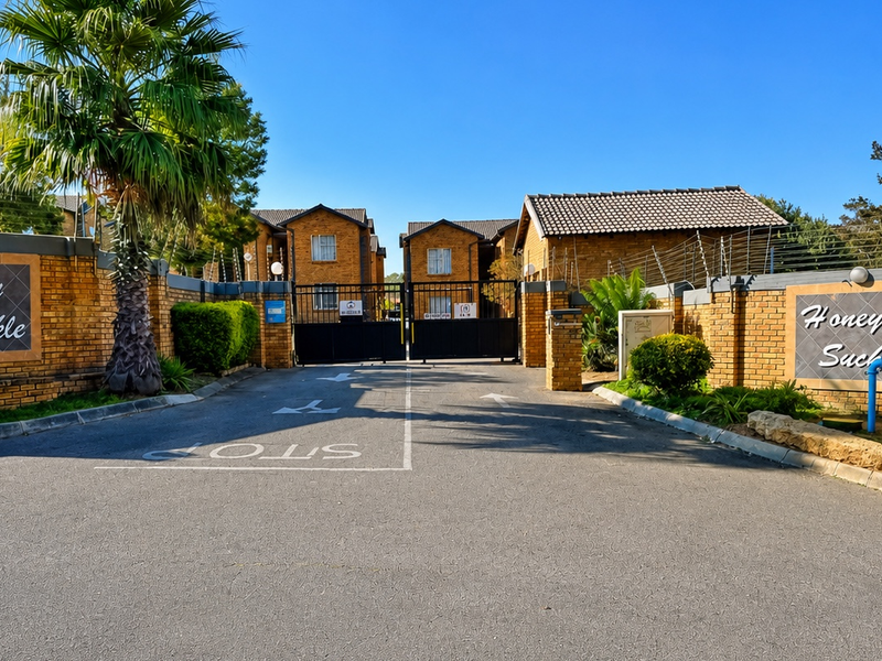 Front entrance view of the Honey Suckle apartment complex, featuring a secure gated access point, brick boundary walls, and established landscaping.