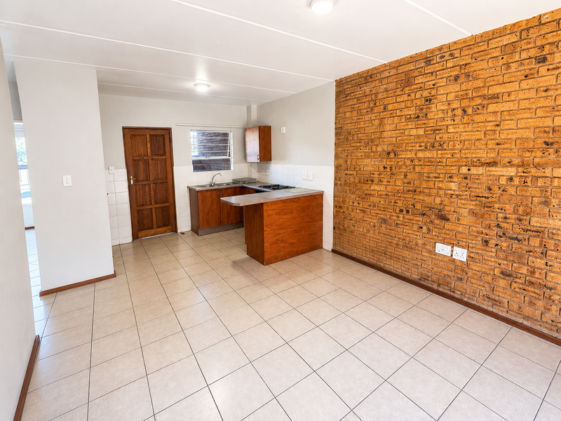 Open-plan living and kitchen area featuring tiled flooring, a prominent exposed brick feature wall, and functional built-in kitchen cabinetry with a hob.