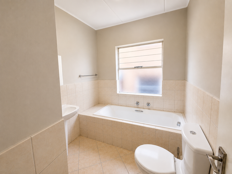 A clean, functional bathroom featuring a built-in bathtub, pedestal basin, toilet, and neutral tiled finishes with a frosted window for privacy.