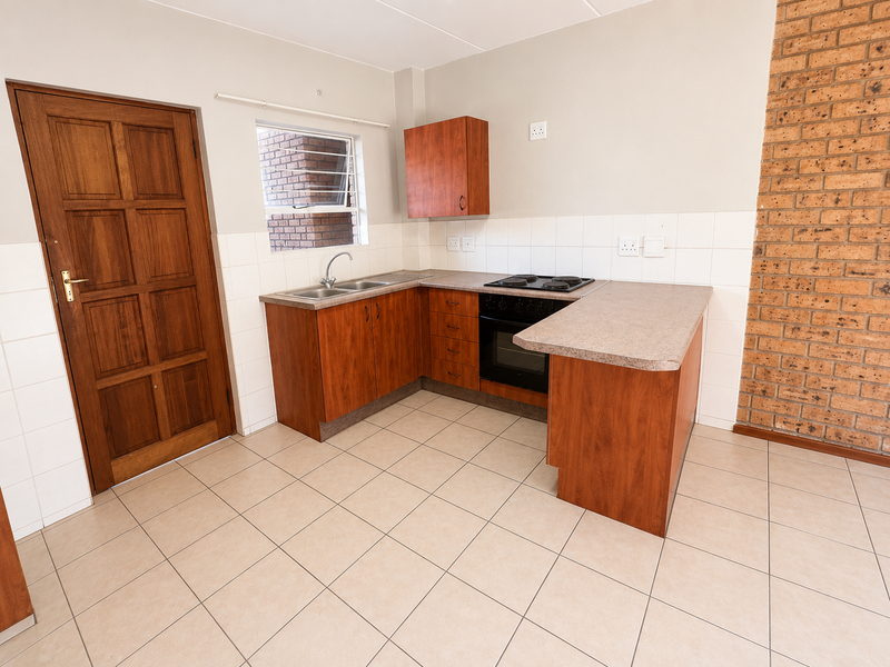 A functional kitchen featuring wood-finish cabinetry, built-in oven and hob, tiled splashbacks, and a distinctive exposed brick feature wall.