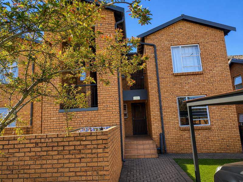 A well-maintained face brick apartment building exterior featuring a paved walkway, secure entrance, and covered carport parking area.