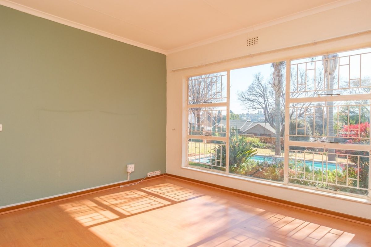 A bright, empty living room featuring laminate flooring, a large window with security bars, and a green accent wall overlooking the garden and pool.