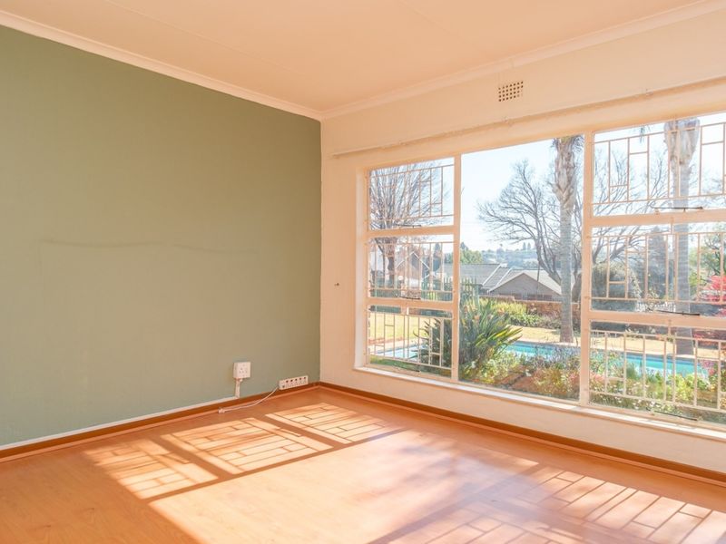 A bright, empty living room featuring laminate flooring, a large window with security bars, and a green accent wall overlooking the garden and pool.