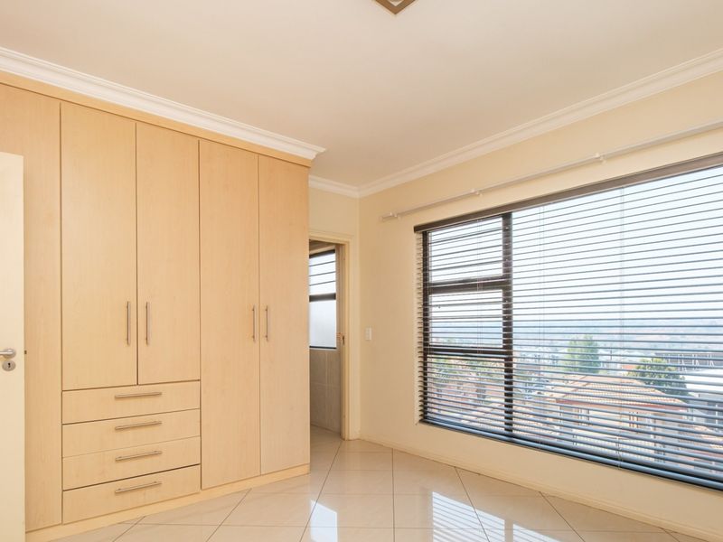 A bright, empty bedroom featuring light wood built-in cupboards, tiled flooring, and large windows with venetian blinds.