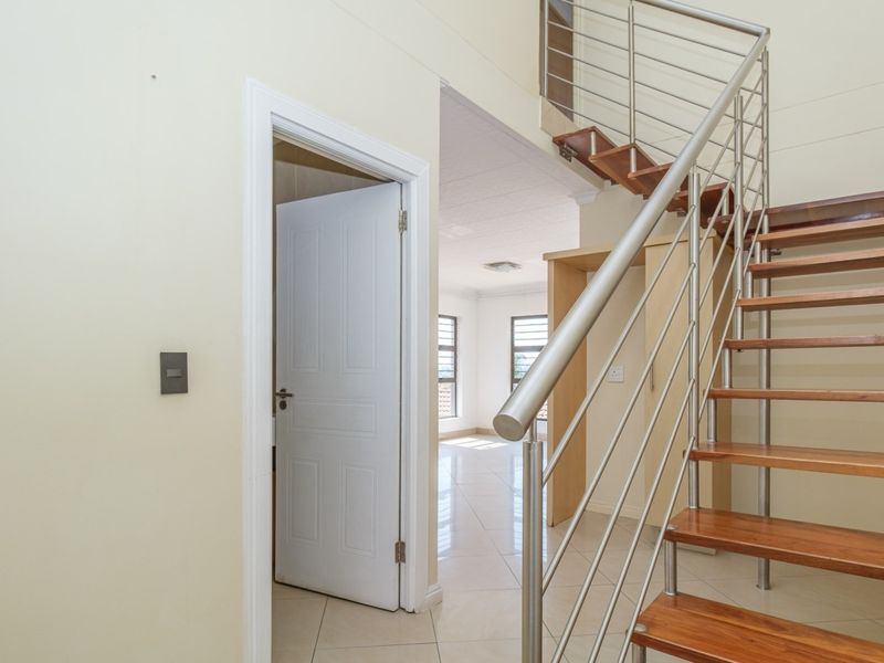 A bright hallway featuring a modern timber staircase with stainless steel balustrades and neutral tiled flooring, leading into an open living area.