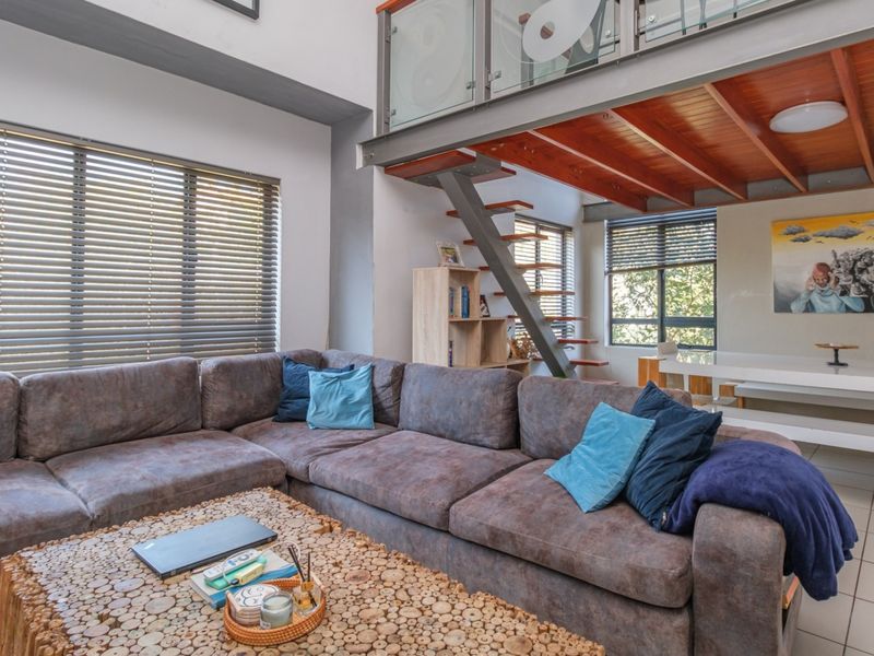 Spacious open-plan living area featuring a mezzanine level with glass balustrade, timber ceiling beams, and large windows with venetian blinds.