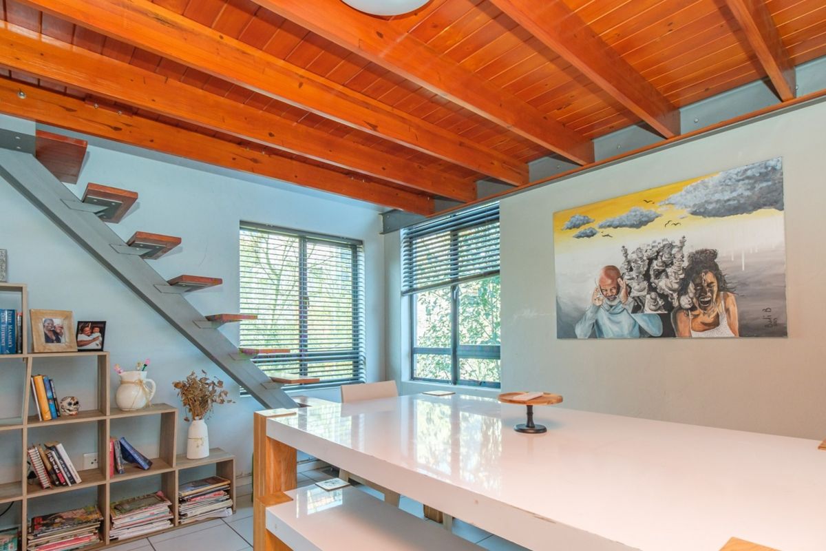 A bright living area featuring a striking exposed timber beam ceiling, a modern floating staircase, and large windows with venetian blinds.