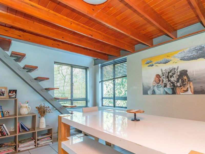 A bright living area featuring a striking exposed timber beam ceiling, a modern floating staircase, and large windows with venetian blinds.
