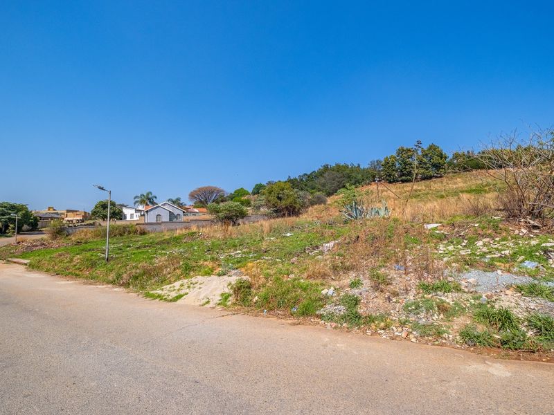 A street-level view of a vacant residential plot featuring a paved road and street lighting, with surrounding neighborhood houses visible in the distance.