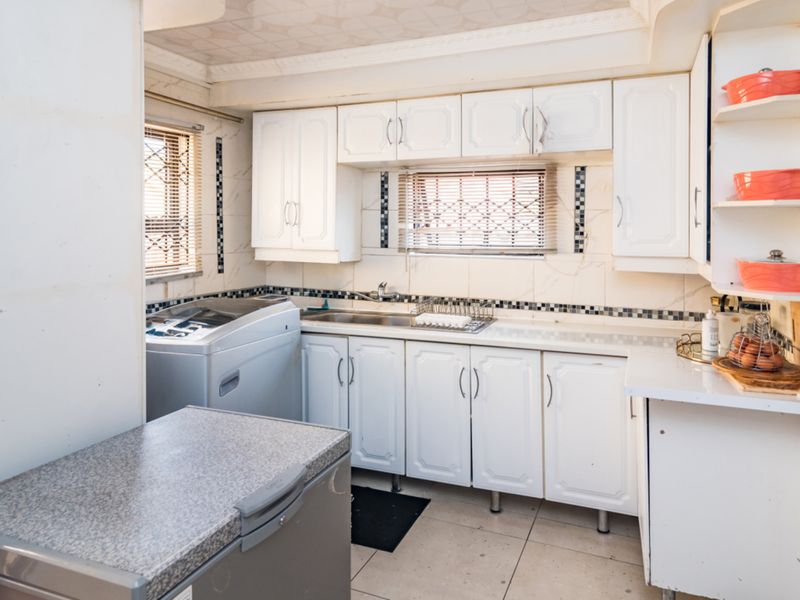 A functional kitchen featuring white built-in cabinetry, tiled backsplash, stainless steel sink, and tiled flooring with security bars on the windows.