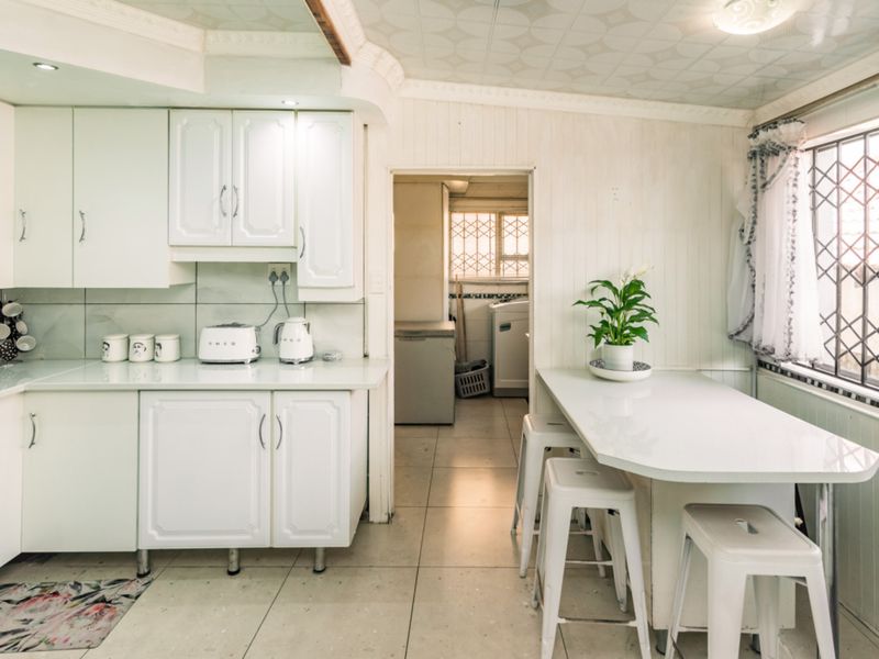A functional kitchen featuring white cabinetry, tiled flooring, and a built-in breakfast bar, with an adjoining scullery area visible through the doorway.