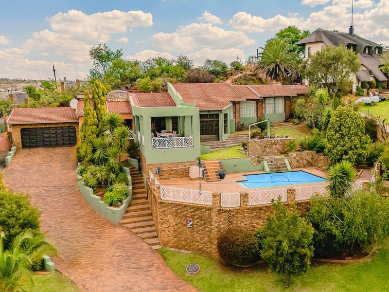 Aerial view of a multi-level residential property featuring a swimming pool, extensive brick paving, a double garage, and established garden landscaping.