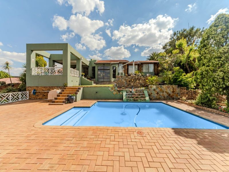 A spacious outdoor pool area featuring a paved patio, a covered patio space, and tiered stone retaining walls leading to the main house structure.