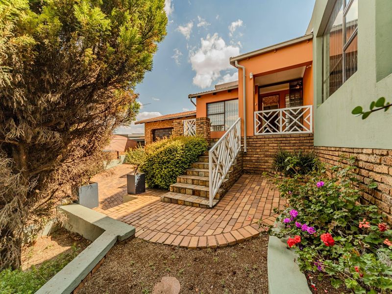 Rear exterior view featuring a brick-paved patio, staircase with metal railings, and established garden beds leading to a covered patio area.