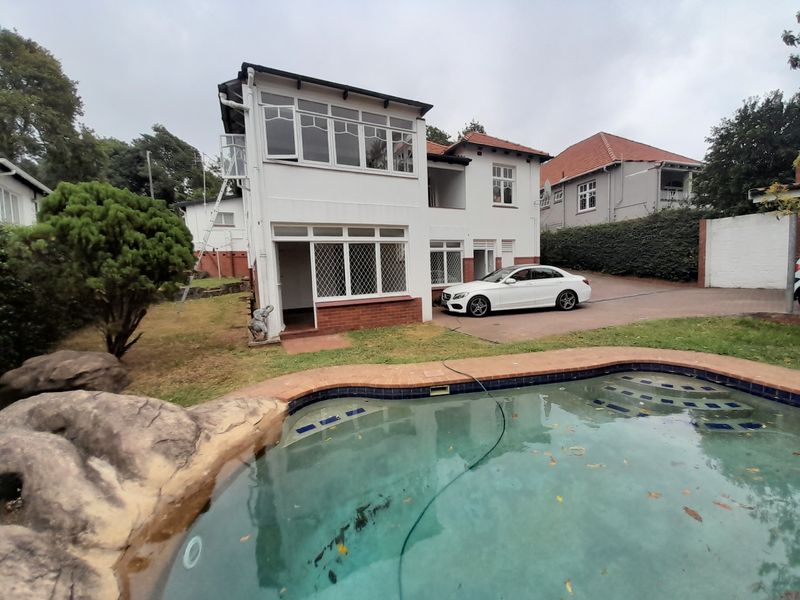 Rear view of a multi-level house featuring a swimming pool with rock landscaping, a paved driveway, and an enclosed upper-level balcony.