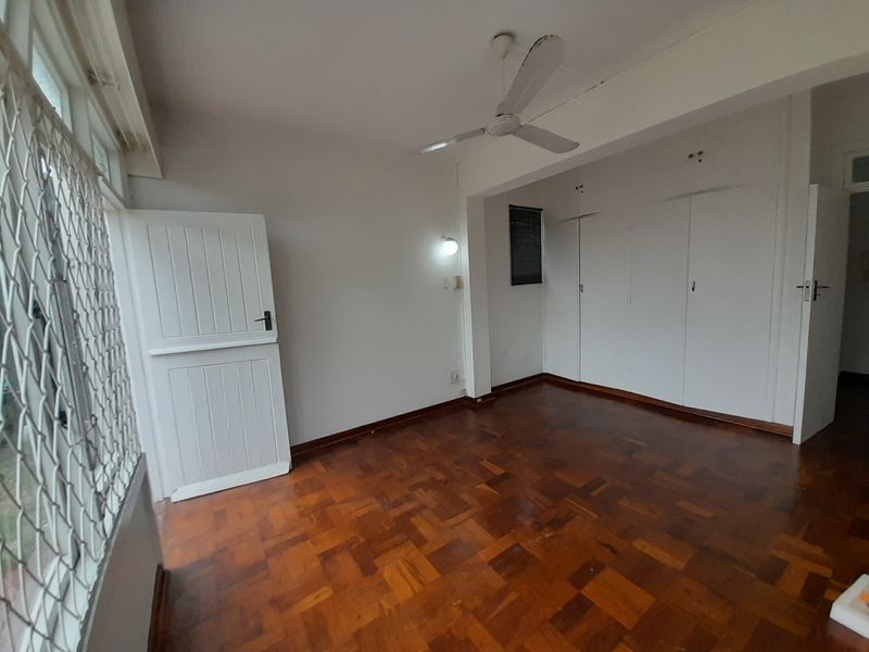 A spacious, empty bedroom featuring classic parquet flooring, built-in storage cupboards, and a ceiling fan, offering a clean canvas for personalization.
