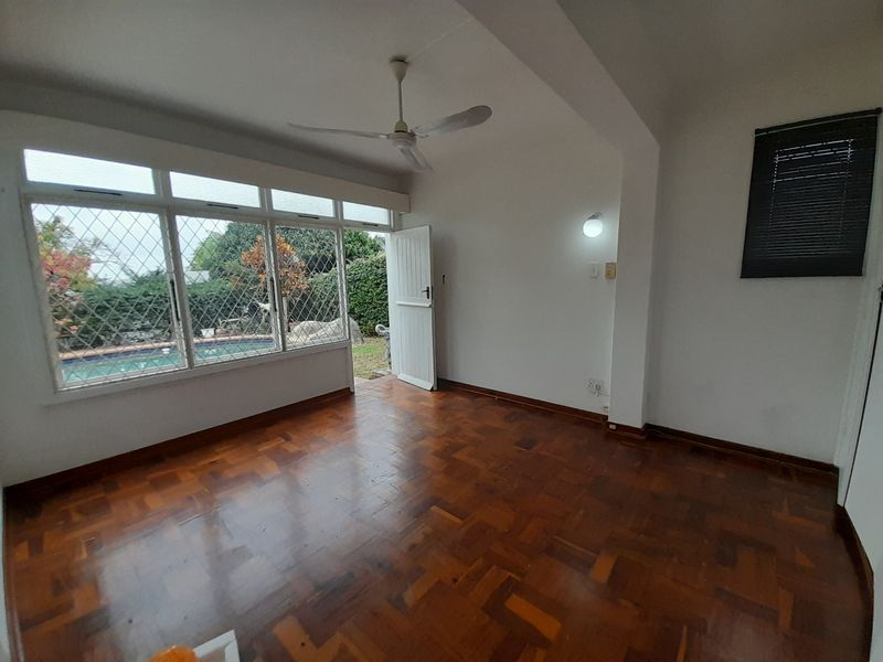 An empty living room featuring classic parquet flooring, a ceiling fan, and large windows with security bars, offering direct access to the outdoor pool area.