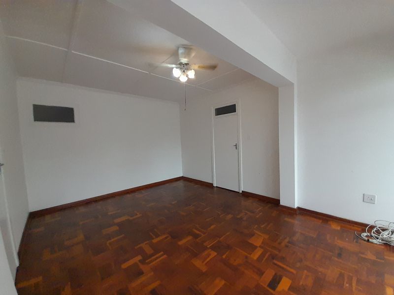 An empty living room featuring polished parquet flooring, a ceiling fan, and neutral white walls, offering a clean and versatile space.