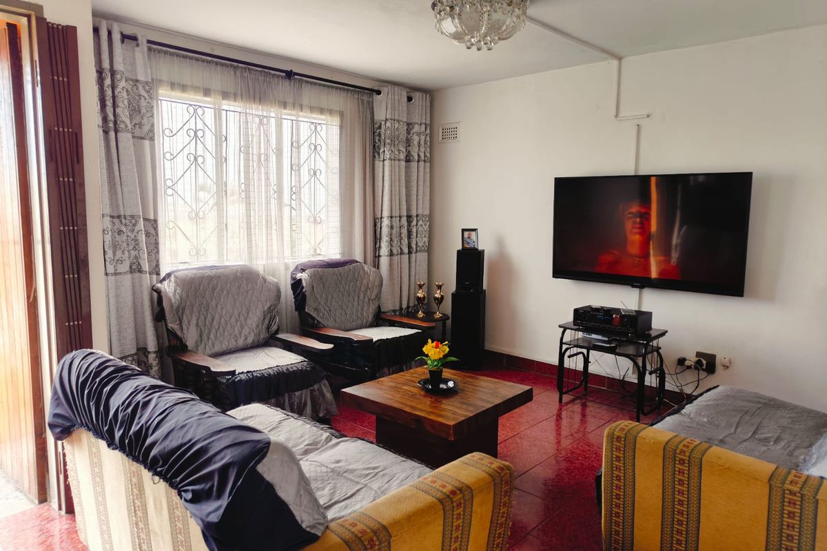 A living room featuring red tiled flooring, a large window with security bars, and a wall-mounted television with surface-mounted electrical wiring.