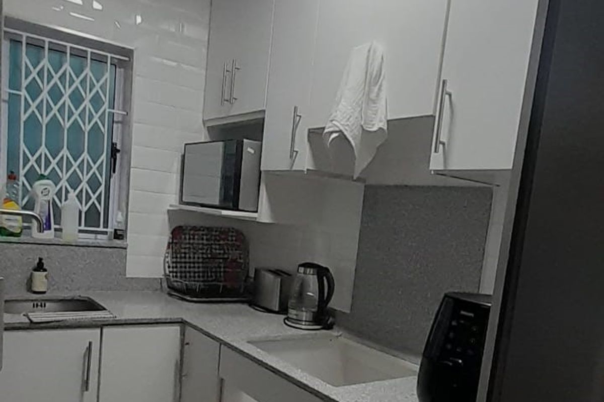 Compact kitchen featuring white cabinetry, stone-look countertops, stainless steel sink, and tiled splashbacks with security bars on the window.