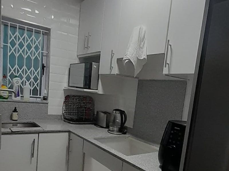 A compact kitchen featuring white cabinetry, laminate countertops, a stainless steel sink, and a tiled backsplash with security bars on the window.