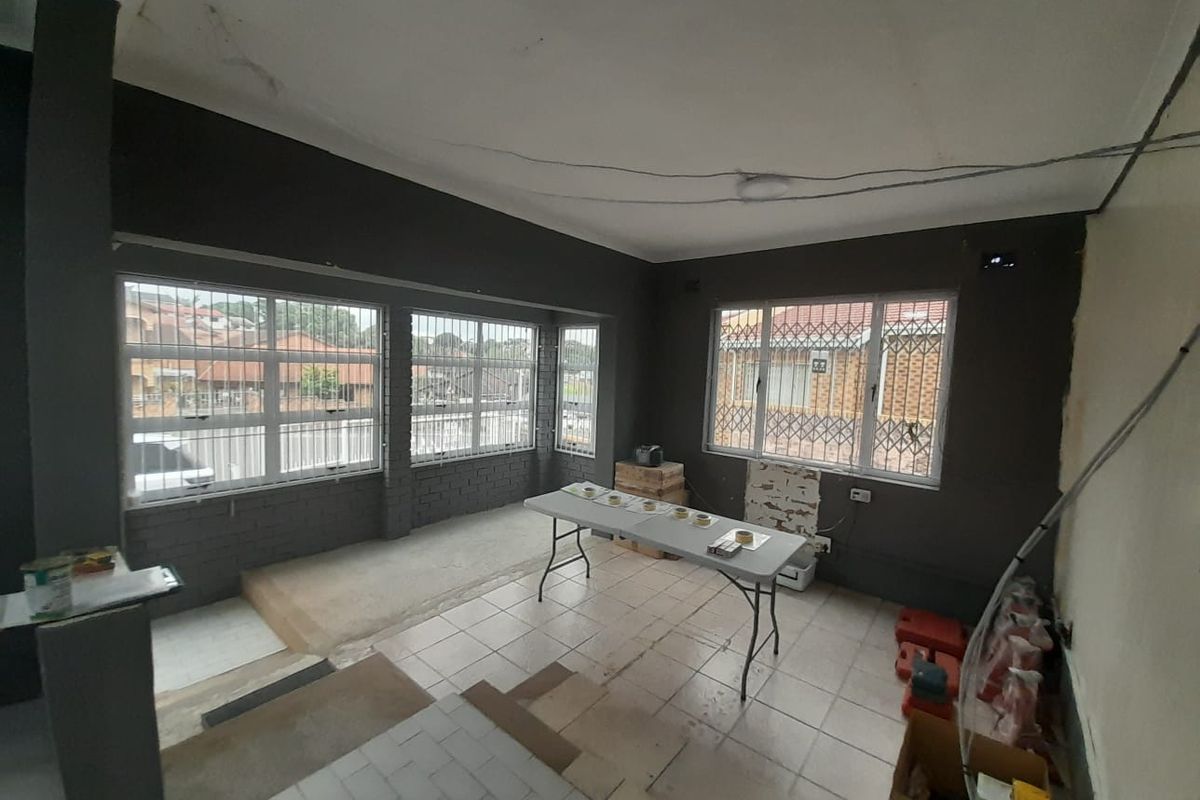 An empty living area featuring tiled floors, large windows with security bars, and exposed wiring, currently in need of renovation and cosmetic repairs.