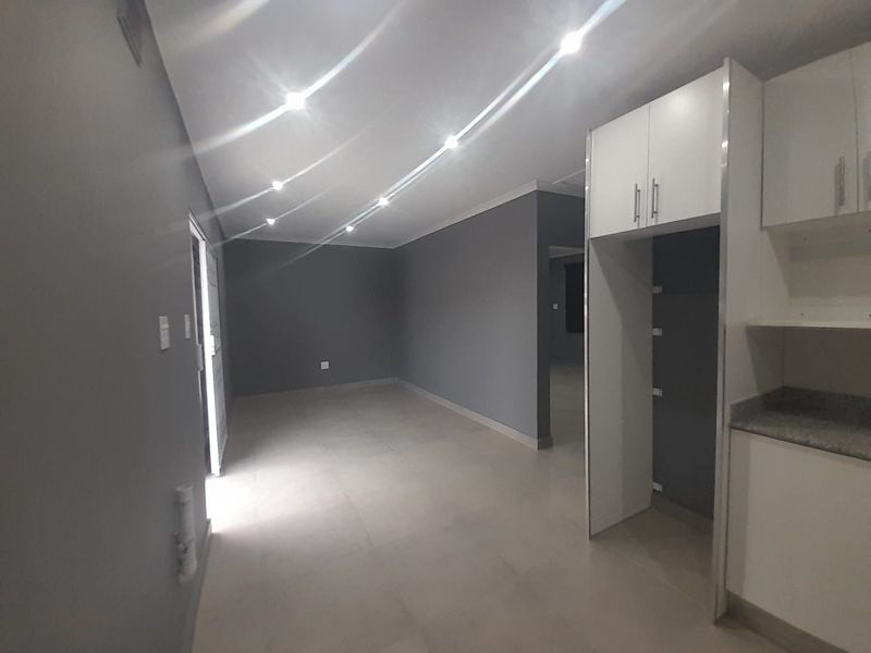 A modern, open-plan living and kitchen area featuring neutral grey walls, tiled flooring, recessed lighting, and clean, white built-in cabinetry.