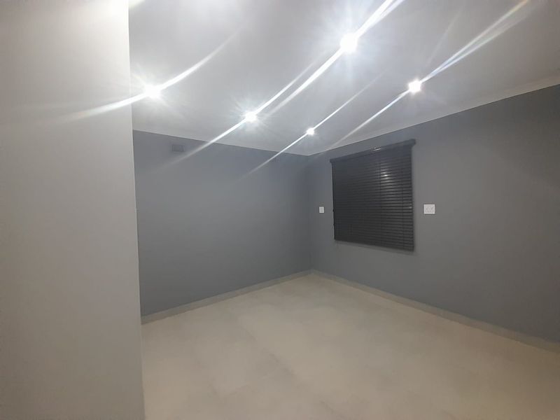 A clean, empty bedroom featuring neutral grey walls, tiled flooring, recessed downlighting, and fitted venetian blinds.